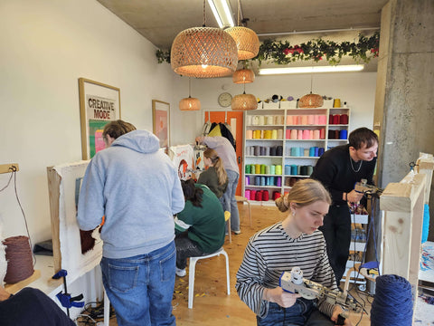 A diverse group of colleagues engaged in a corporate team-building activity, happily tufting their custom designs together at large frames in a modern, light-filled studio.