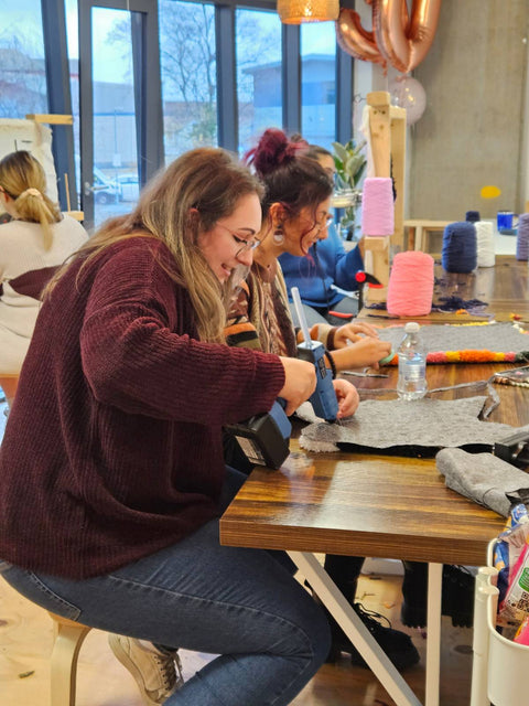 Two women sitting at a table with craft materials, smiling and engaged in an rug making activity.