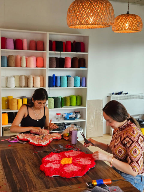 Two women working on a rug in a room with colorful yarn on shelves.