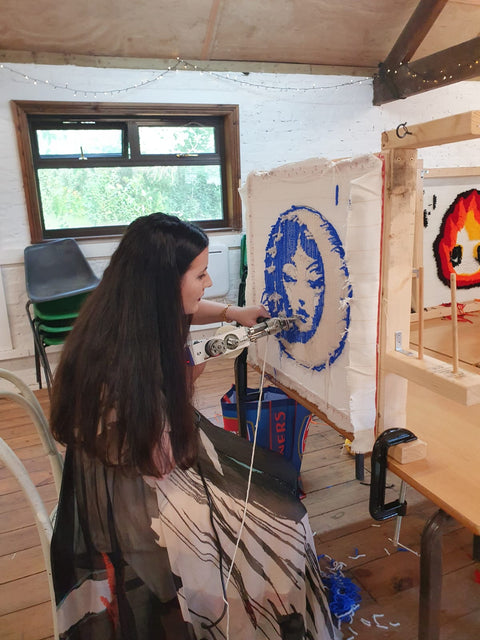 Women using a tufting gun to paint on a large monk cloth canvas in an indoor setting.